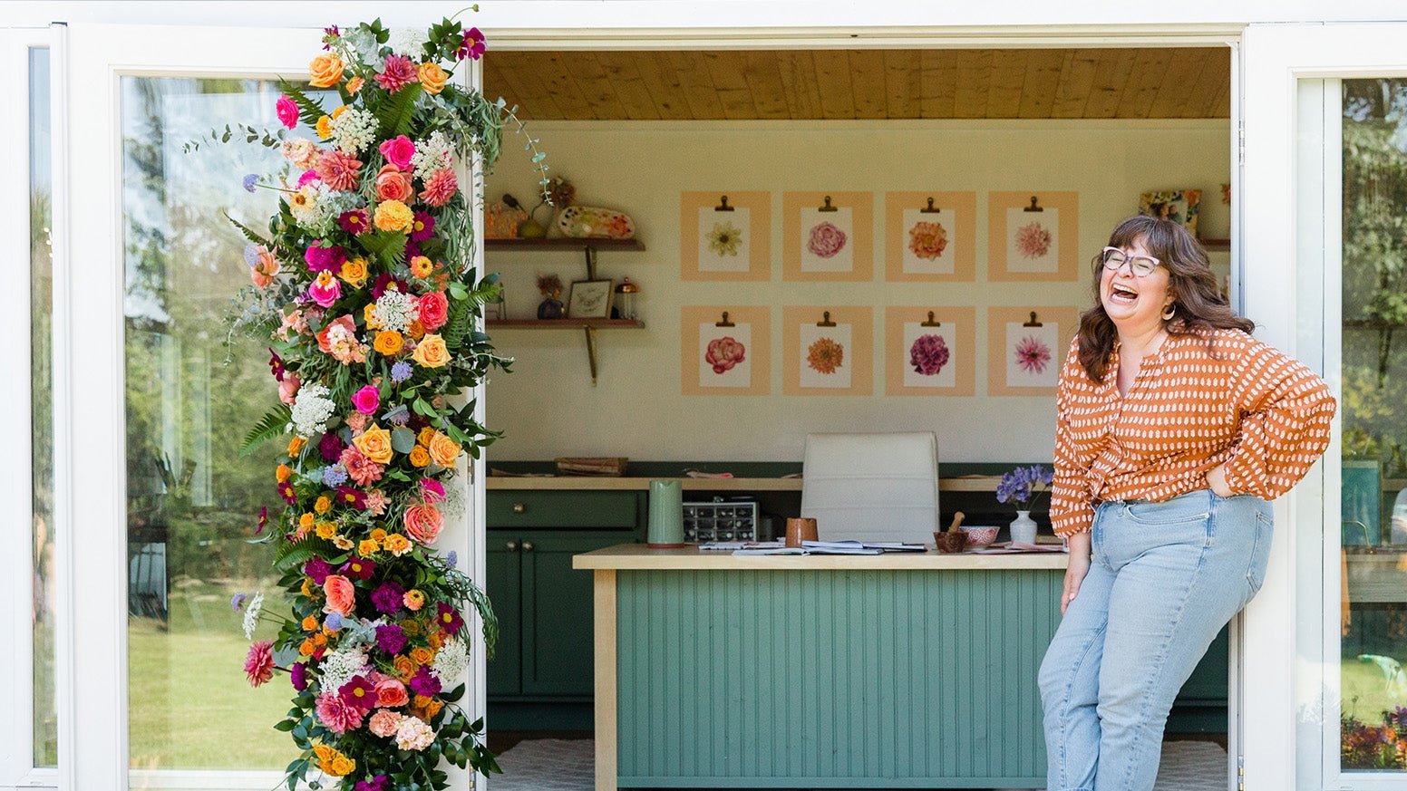 Kate laughing at the door of her shed with watercolor paintings and a desk in the background and a big wreath of flowers on the door.